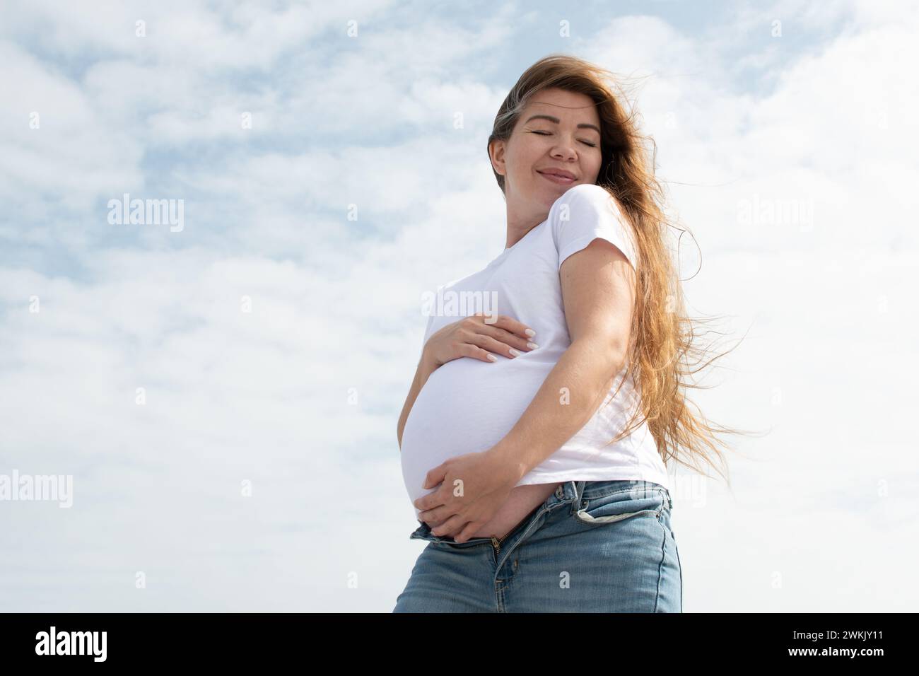 Heureuse femme enceinte caucasienne tenant son ventre sur le fond du ciel. t-shirt blanc et jeans. Troisième trimestre. Concept de grossesse heureuse Banque D'Images