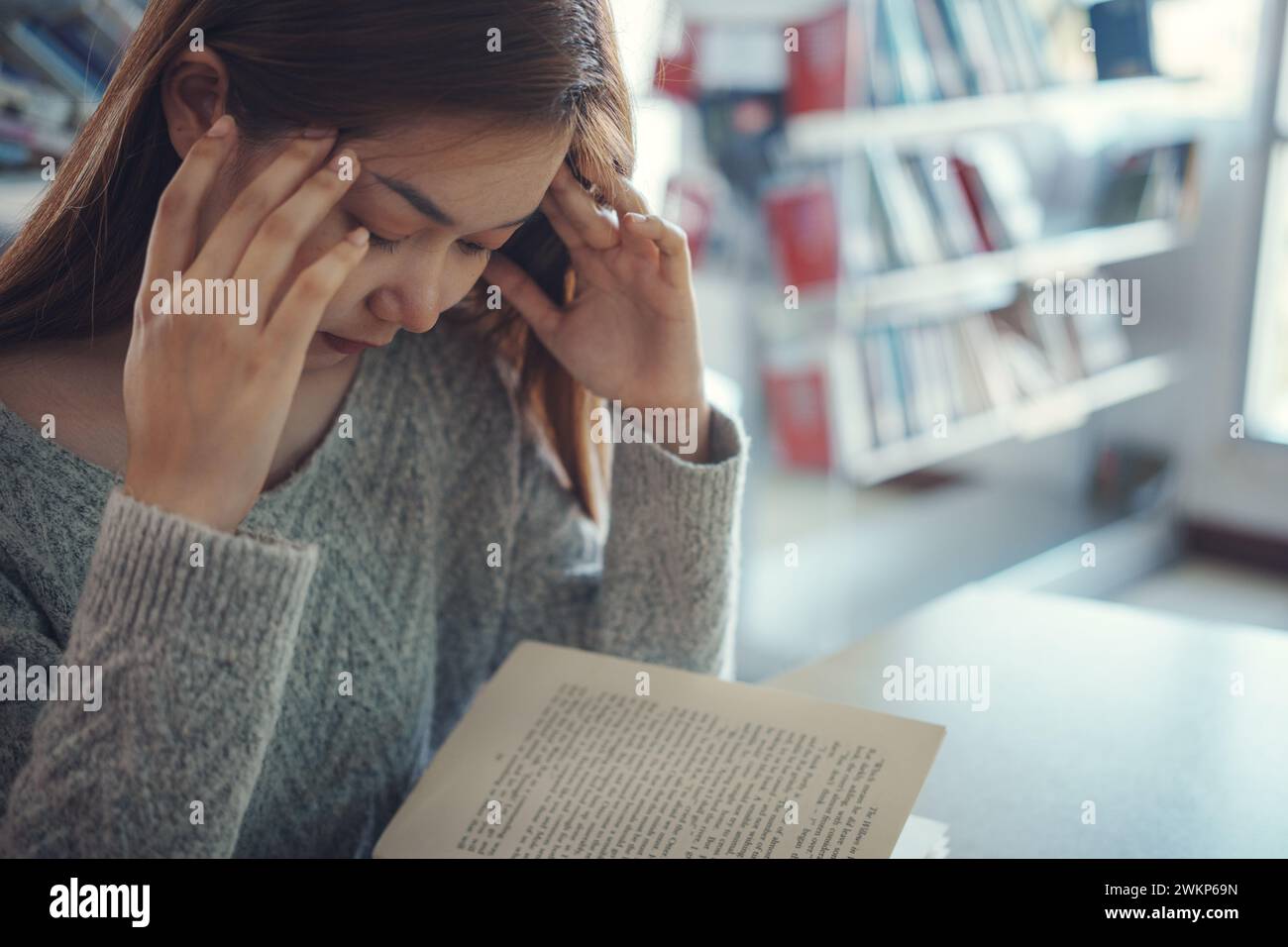 Jeune étudiante stressée assise à une table en bois et lisant un livre pendant la préparation de l'examen à la bibliothèque. Banque D'Images