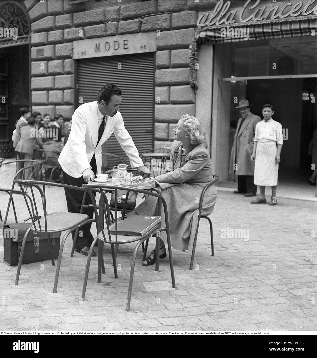 Vaction en Italie dans les années 1950 Une jeune femme assise à une table de café dans une rue animée de Milan. Un serveur se tient à côté d'elle étant poli et charmé par la jeune fille blonde suédoise. Milan Italie 1950. Photo Kristoffersson Ref DV19 *** local Caption *** © Classic Picture Library. Tous droits réservés. Protégé par une signature numérique. La surveillance et la protection de l'image sont activées sur cette image. La licence ; présentation ou newsletter N'inclut PAS l'utilisation sur les médias sociaux. Banque D'Images