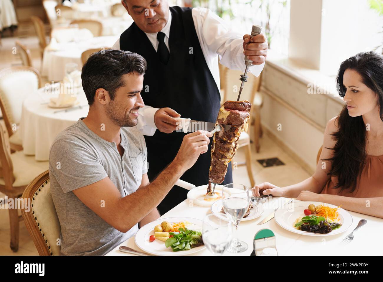 Couple heureux, date et serveur avec kebab pour servir, dîner fin ou dîner romantique à table. Jeune homme et femme avec chef coupant brochette de nourriture Banque D'Images
