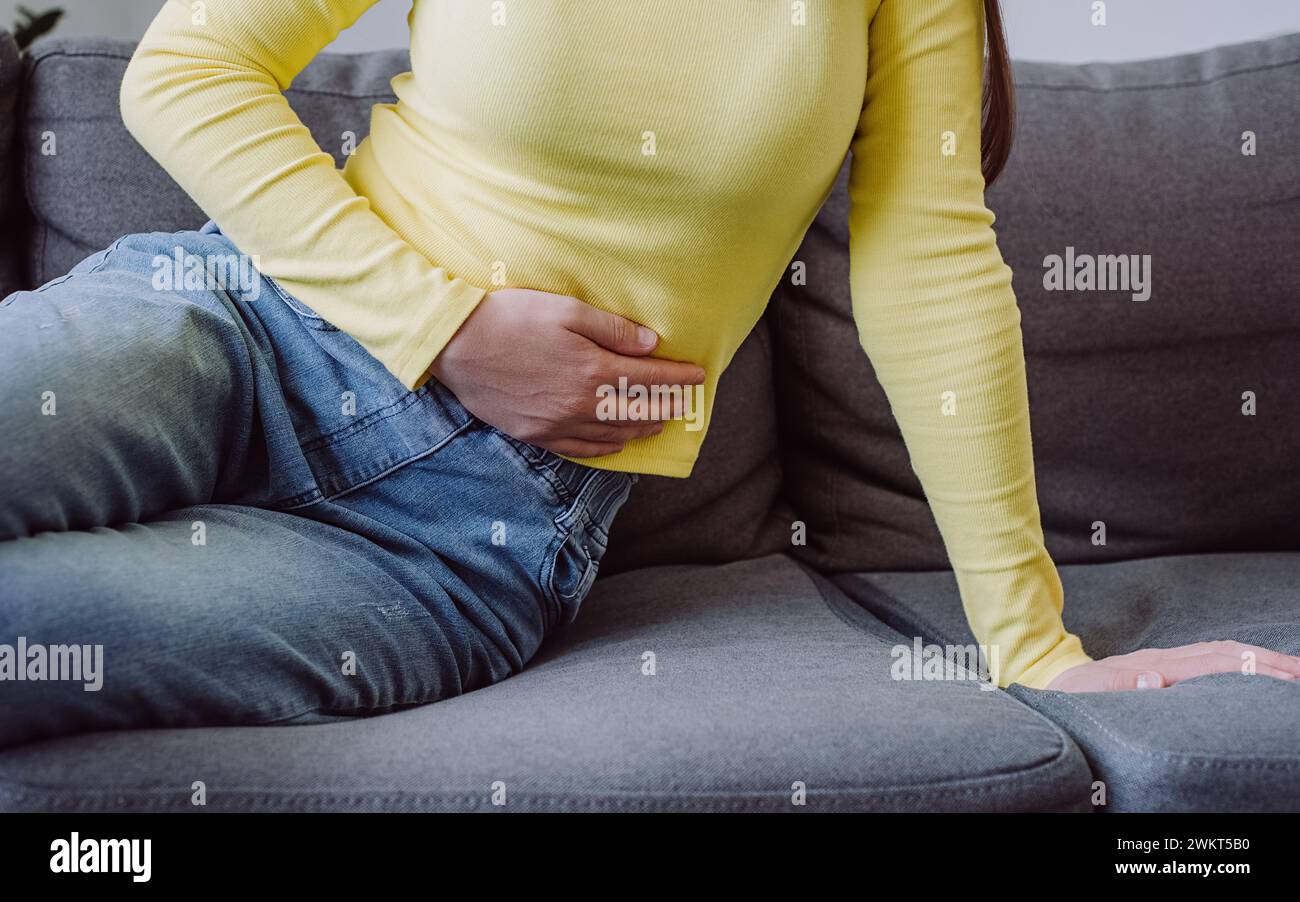 Gros plan de jeune femme inconnue tenant le ventre assis sur le canapé à la maison, se sentant inconfortable, concept de problème de santé, souffrant de maux d'estomac, intoxication alimentaire Banque D'Images