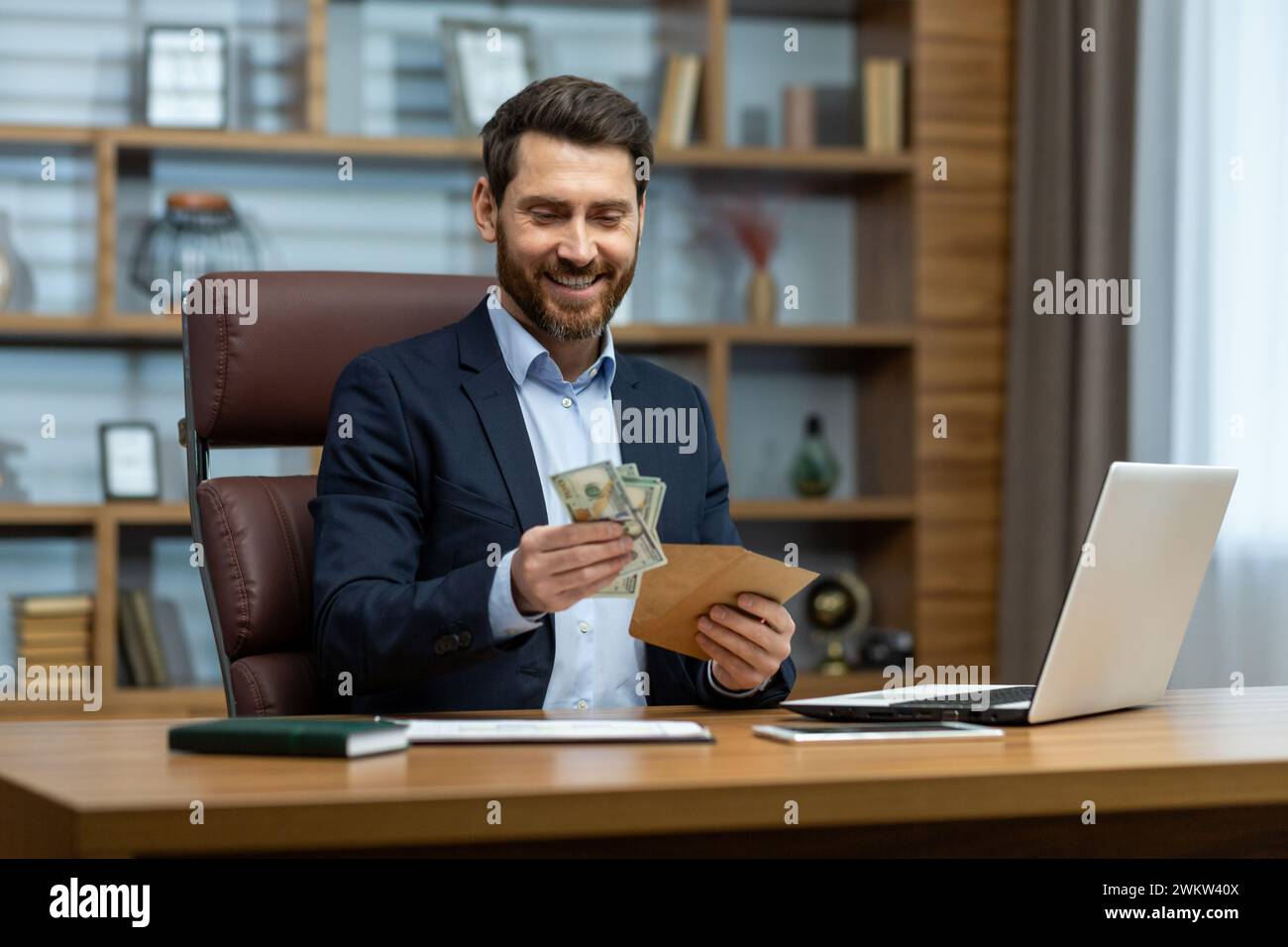 Photo d'un jeune homme d'affaires heureux assis à un bureau dans le bureau et tenant une enveloppe avec de l'argent liquide dans ses mains, regardant satisfait de la b reçue Banque D'Images