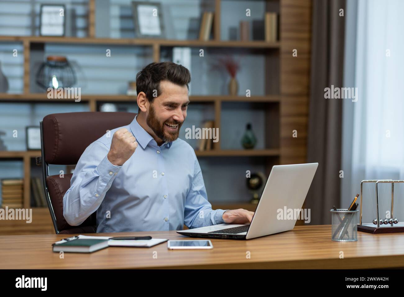 Heureux jeune homme d'affaires assis dans le bureau au bureau avec un ordinateur portable et se réjouit du succès, en regardant la fenêtre, montrant une gestu de victoire Banque D'Images