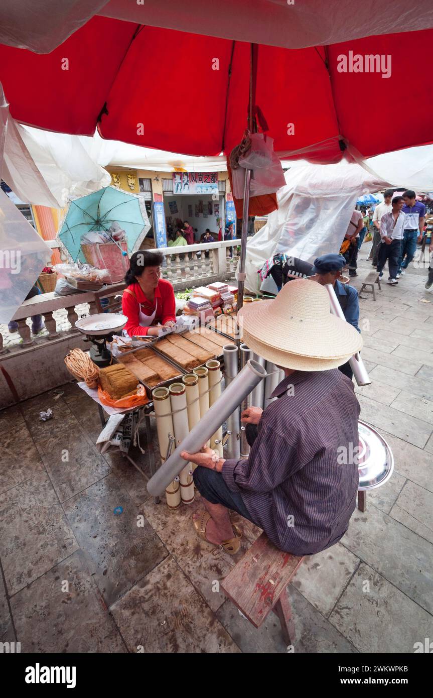 Un homme à un étal de vendeurs de tabac dans un marché chinois du Yunnan fumant une pipe commerciale. Banque D'Images