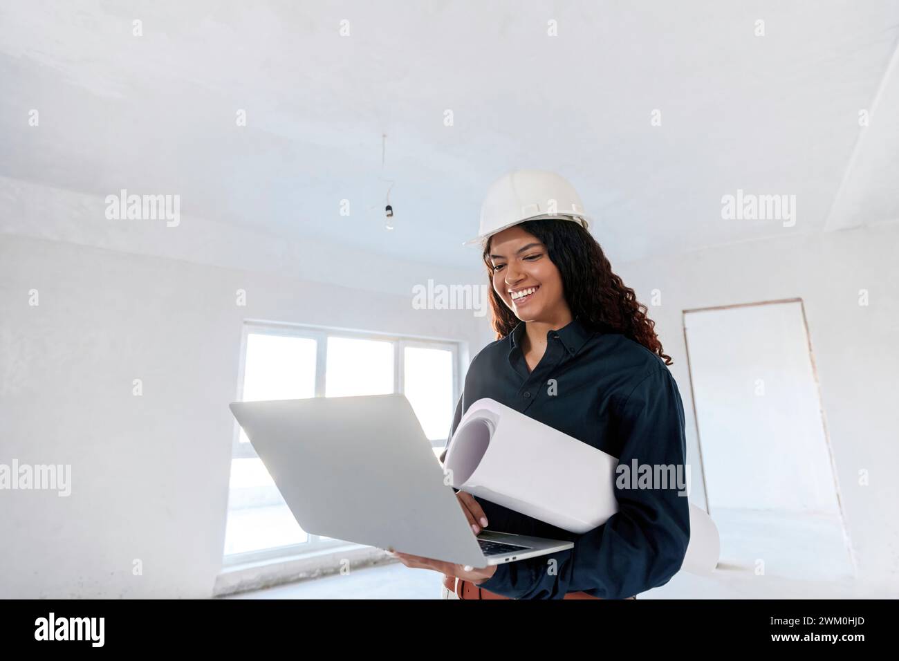 Heureux jeune architecte portant un casque dur tenant le plan et utilisant un ordinateur portable sur le site Banque D'Images