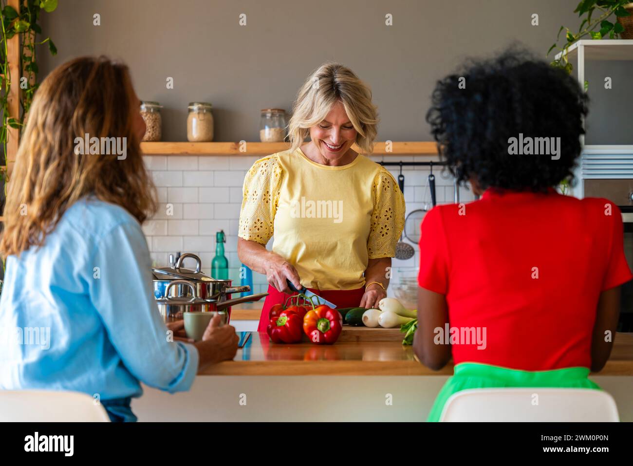 Femme souriante préparant la nourriture près d'amis assis dans la cuisine à la maison Banque D'Images