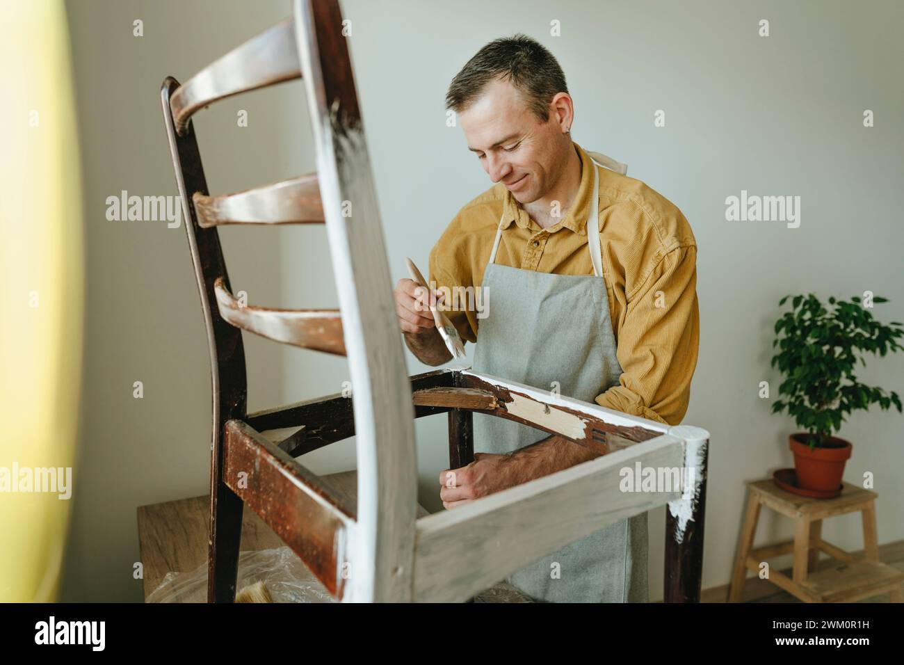 Homme souriant portant tablier et chaise de coloriage avec brosse à la maison Banque D'Images
