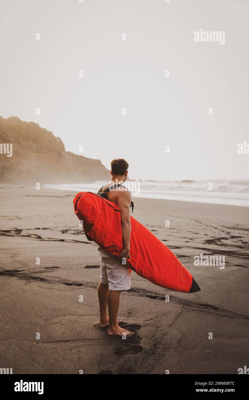 Jeune homme portant une planche de surf et debout sur la plage Banque D'Images