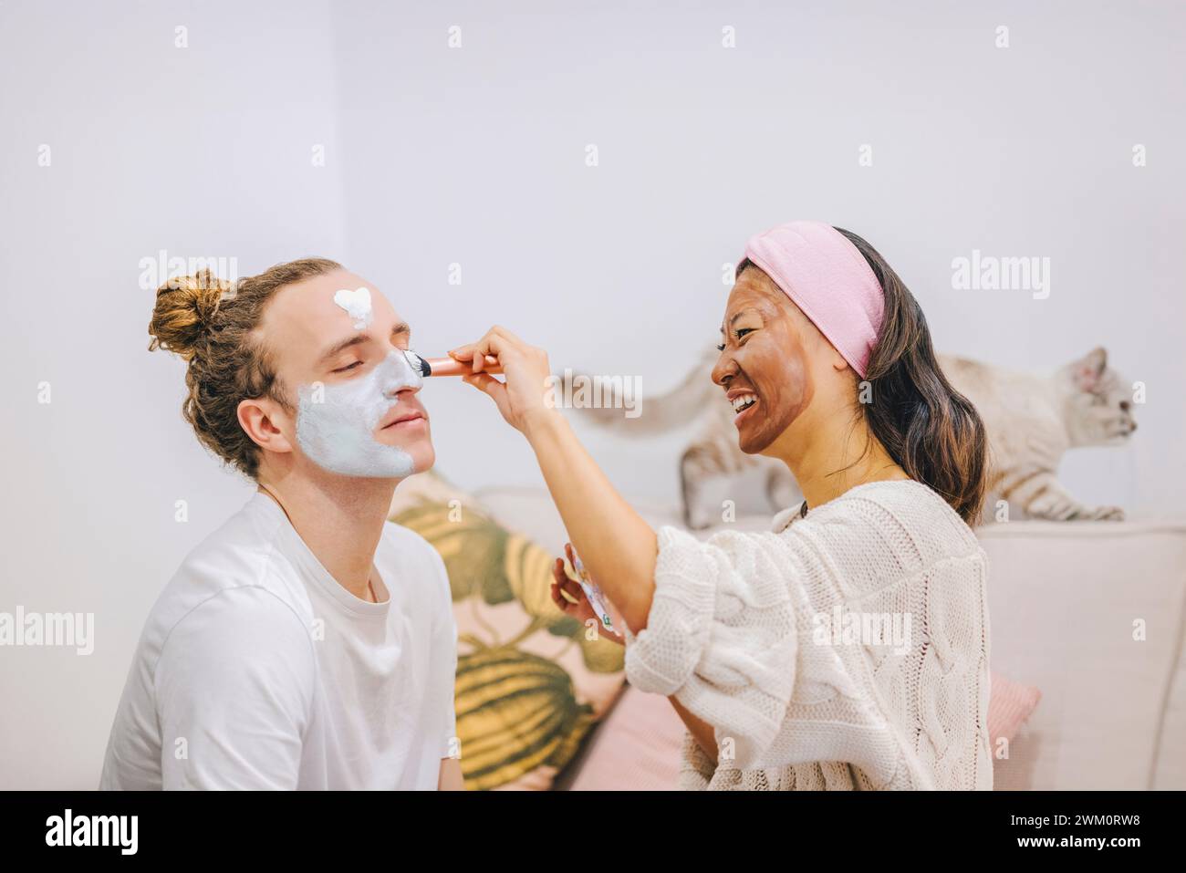 Femme heureuse appliquant le masque de soin de la peau sur le visage de petit ami à la maison Banque D'Images
