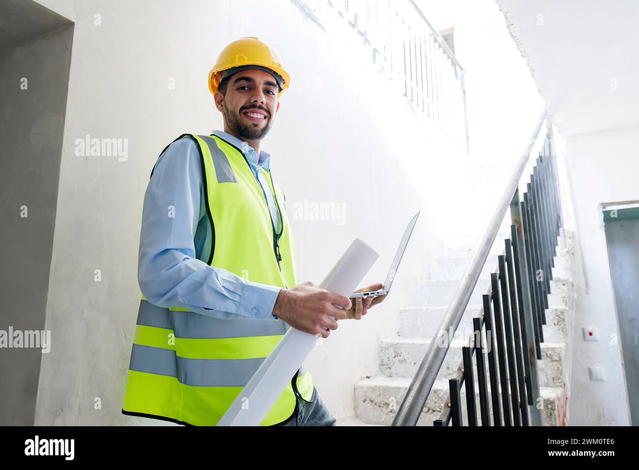 Architecte souriant portant un casque et debout sur les marches tenant l'ordinateur portable sur le site Banque D'Images