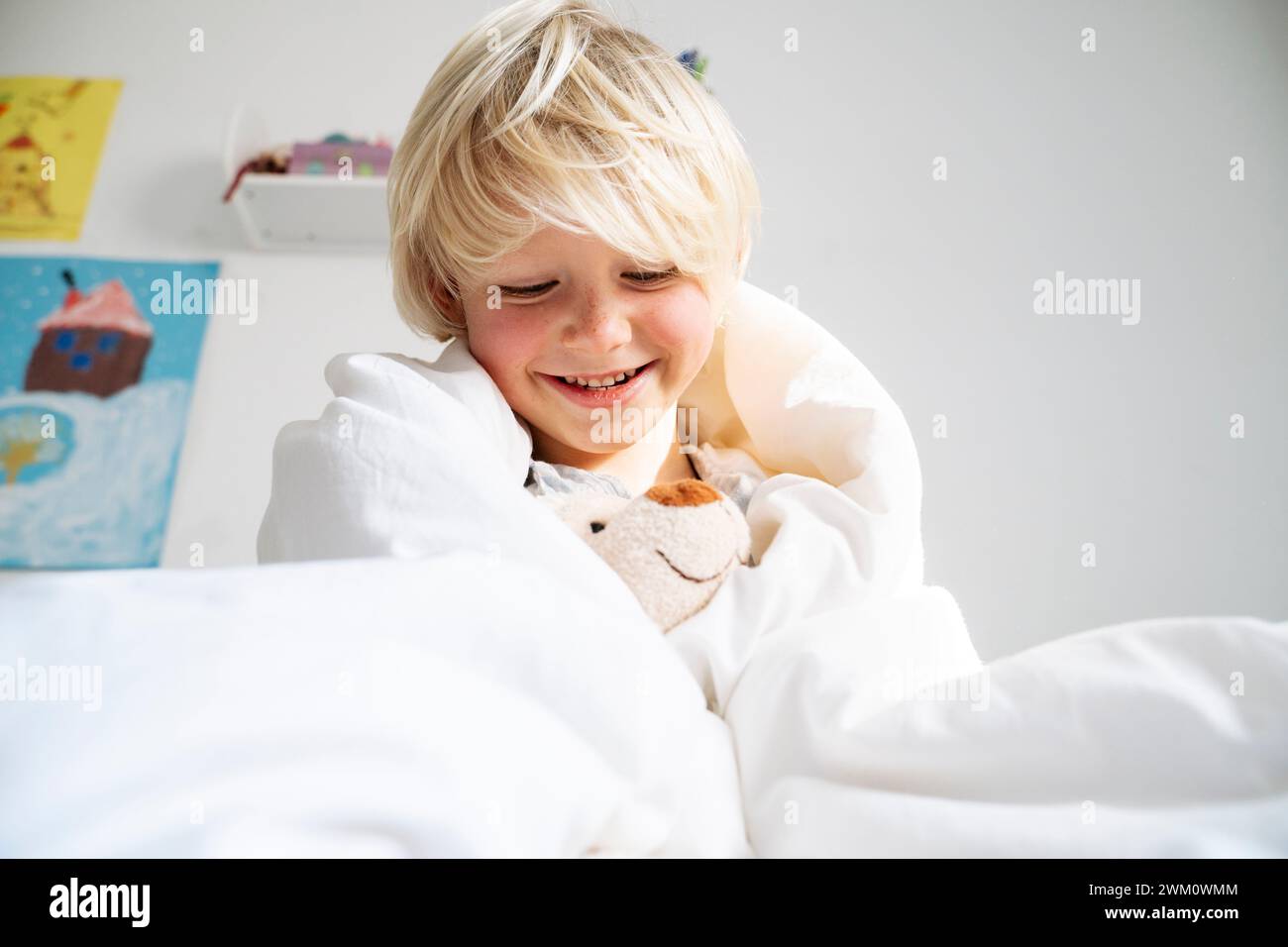 Garçon heureux jouant avec un ours en peluche enveloppé dans une couverture à la maison Banque D'Images