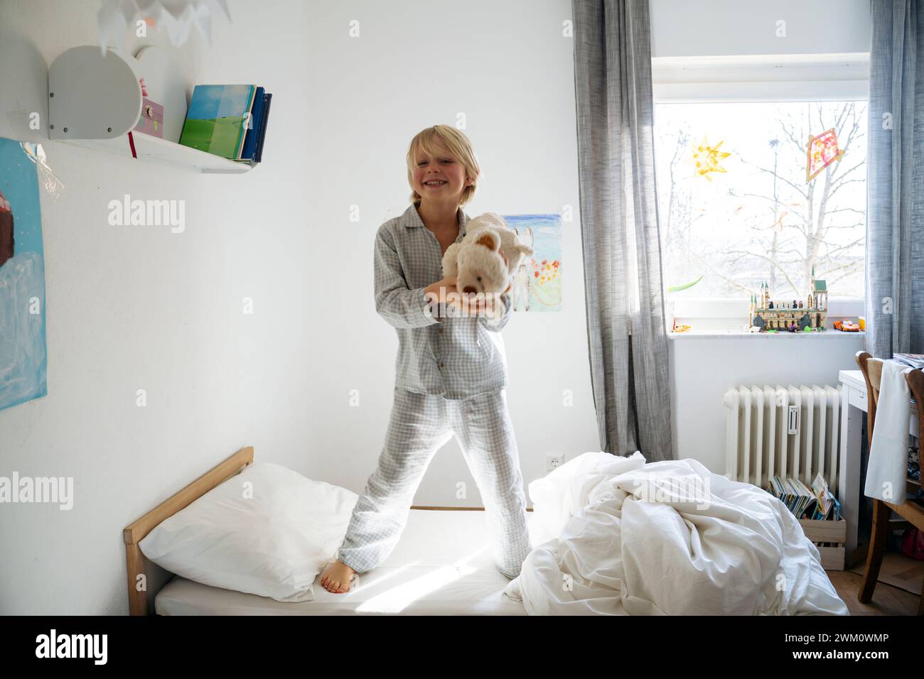 Garçon heureux tenant un ours en peluche et debout sur le lit à la maison Banque D'Images