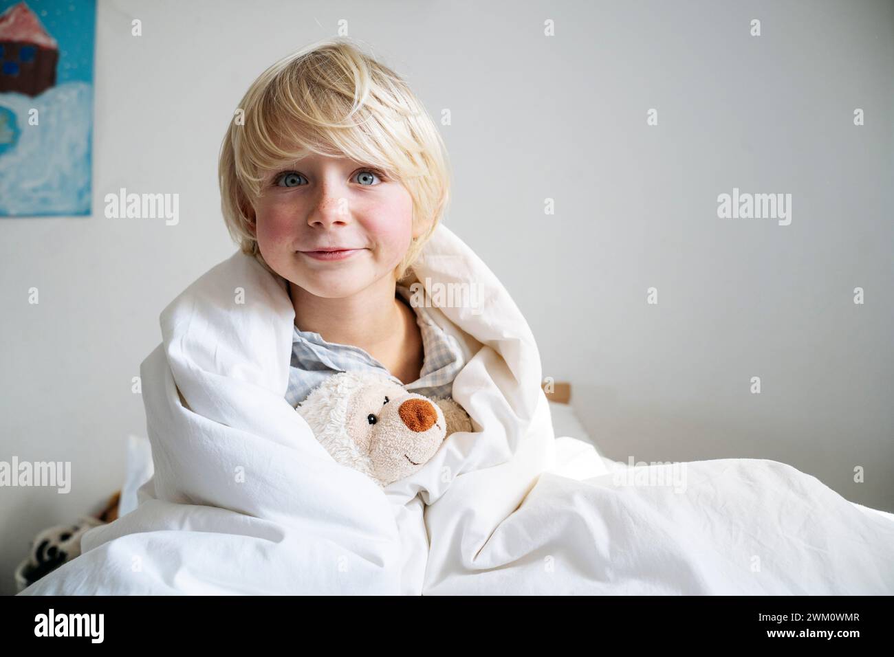 Garçon blond avec ours en peluche enveloppé dans une couverture à la maison Banque D'Images