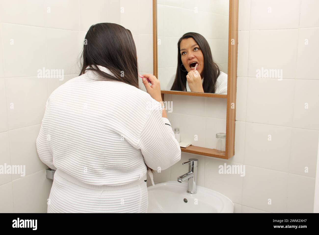 Latina femme de 40 ans se brosse et prend soin de ses dents pour prévenir les caries habillées en peignoir dans la salle de bain devant le miroir Banque D'Images