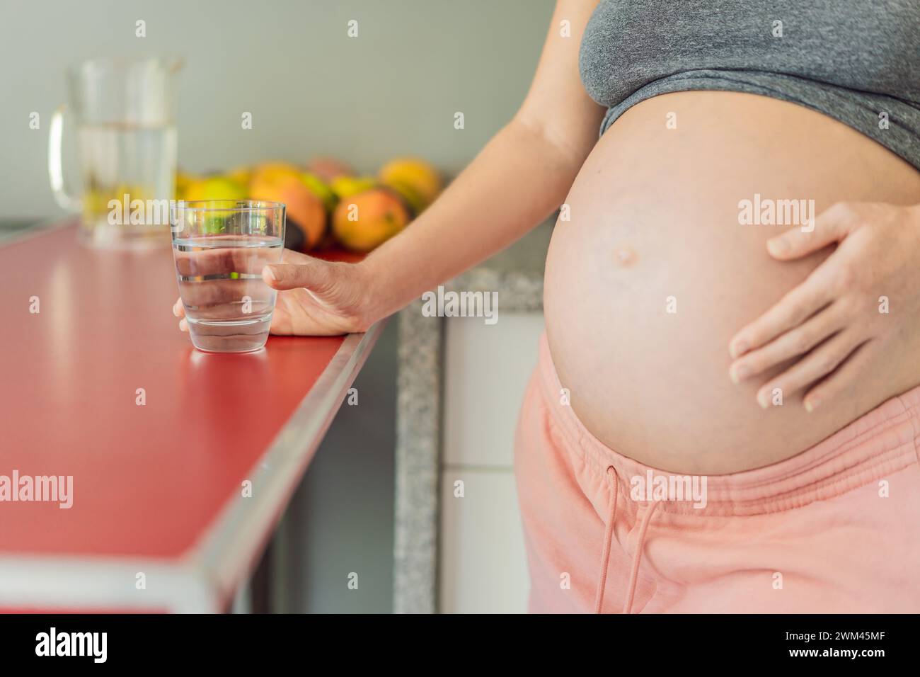 Embrassant les bienfaits vitaux de l'eau pendant la grossesse, une femme enceinte se tient dans la cuisine avec un verre, soulignant le rôle crucial de l'hydratation Banque D'Images