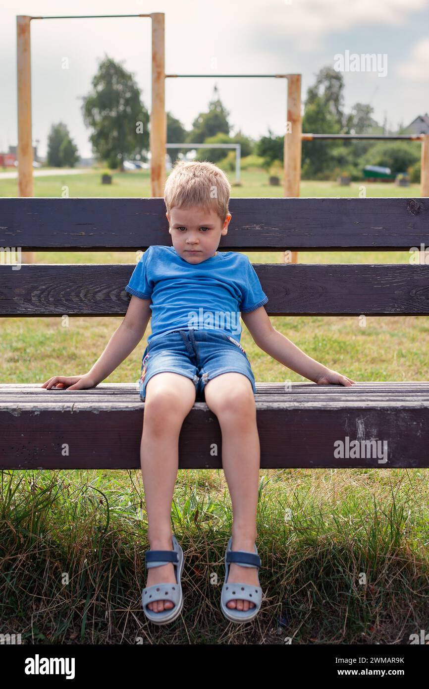 Un petit garçon triste est assis sur un banc dans une aire de jeux extérieure. Humeur triste dans le concept de dépression infantile Banque D'Images