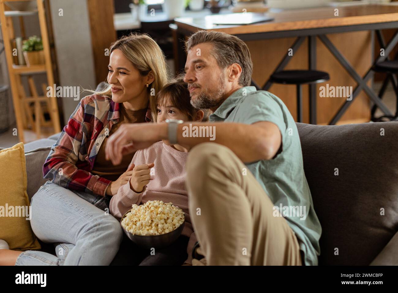 Une famille de trois est confortablement nichée sur un canapé, leurs visages reflétant l'excitation et l'attention comme ils partagent un bol de pop-corn pendant un susp Banque D'Images