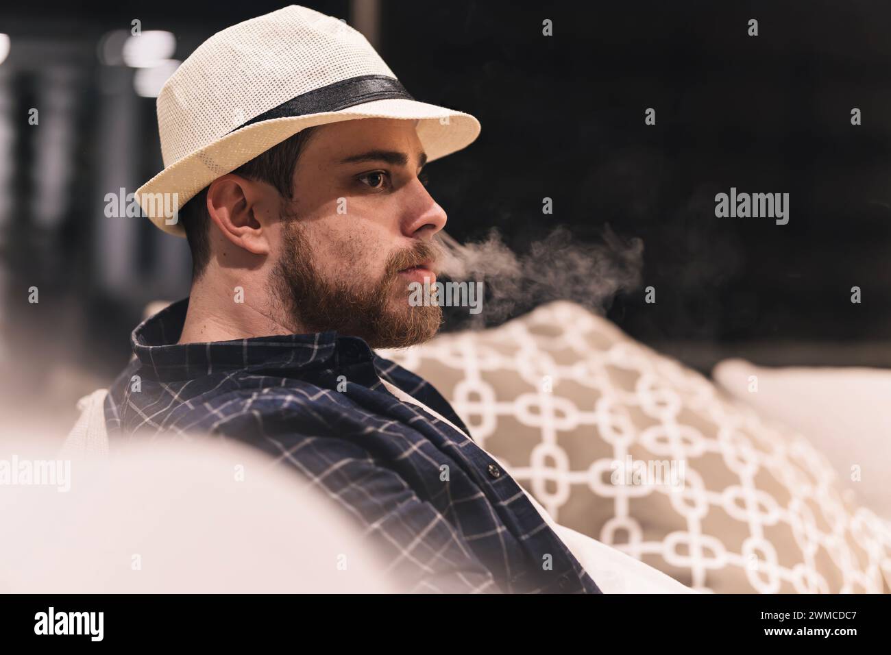 Jeune homme dans le chapeau soufflant de la fumée assis sur le canapé à la maison après le retour du travail. Concept de style de vie. Copier l'espace Banque D'Images