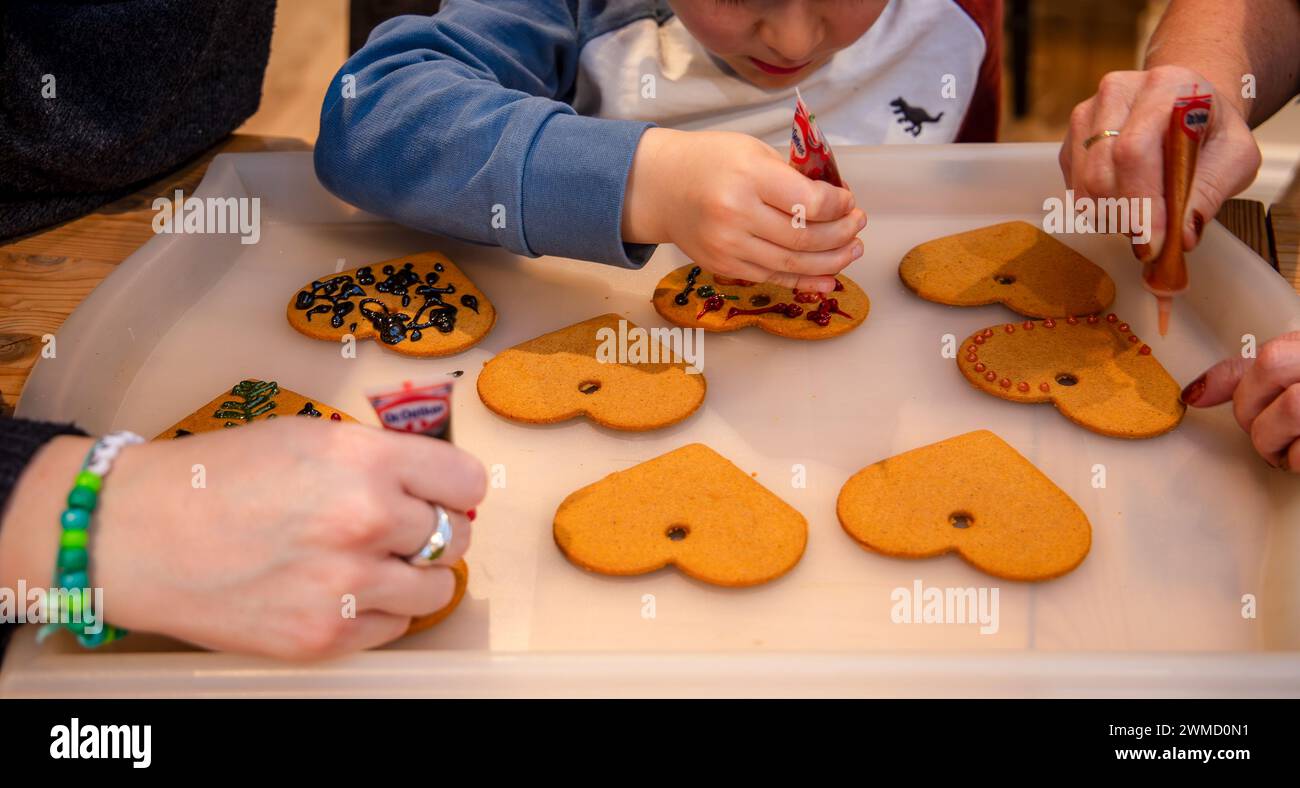 Jeune enfant de 5 ans décorant des biscuits en forme de coeur en pain d'épices pour Noël avec l'aide des adultes Banque D'Images