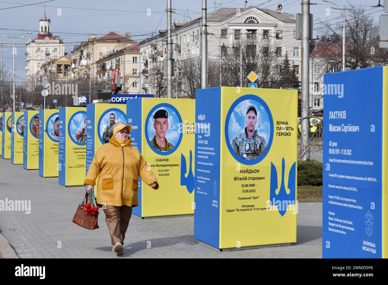 Une femme âgée passe devant des stands de portraits de soldats ukrainiens tombés au combat dans le centre de Zaporizhzhia. Le président ukrainien Volodymyr Zelensky a marqué le deuxième anniversaire de l'invasion russe en déclarant que son pays "réussira". L'Ukraine a défié les prédictions de combattre la Russie, mais il y a des signes que la guerre tourne en faveur de la Russie. Le gouvernement ukrainien a cessé de partager le nombre de ses militaires morts il y a longtemps. Mais le nombre toujours croissant de tombes fraîches, des deux côtés des lignes de front, dresse un tableau douloureusement clair. Banque D'Images