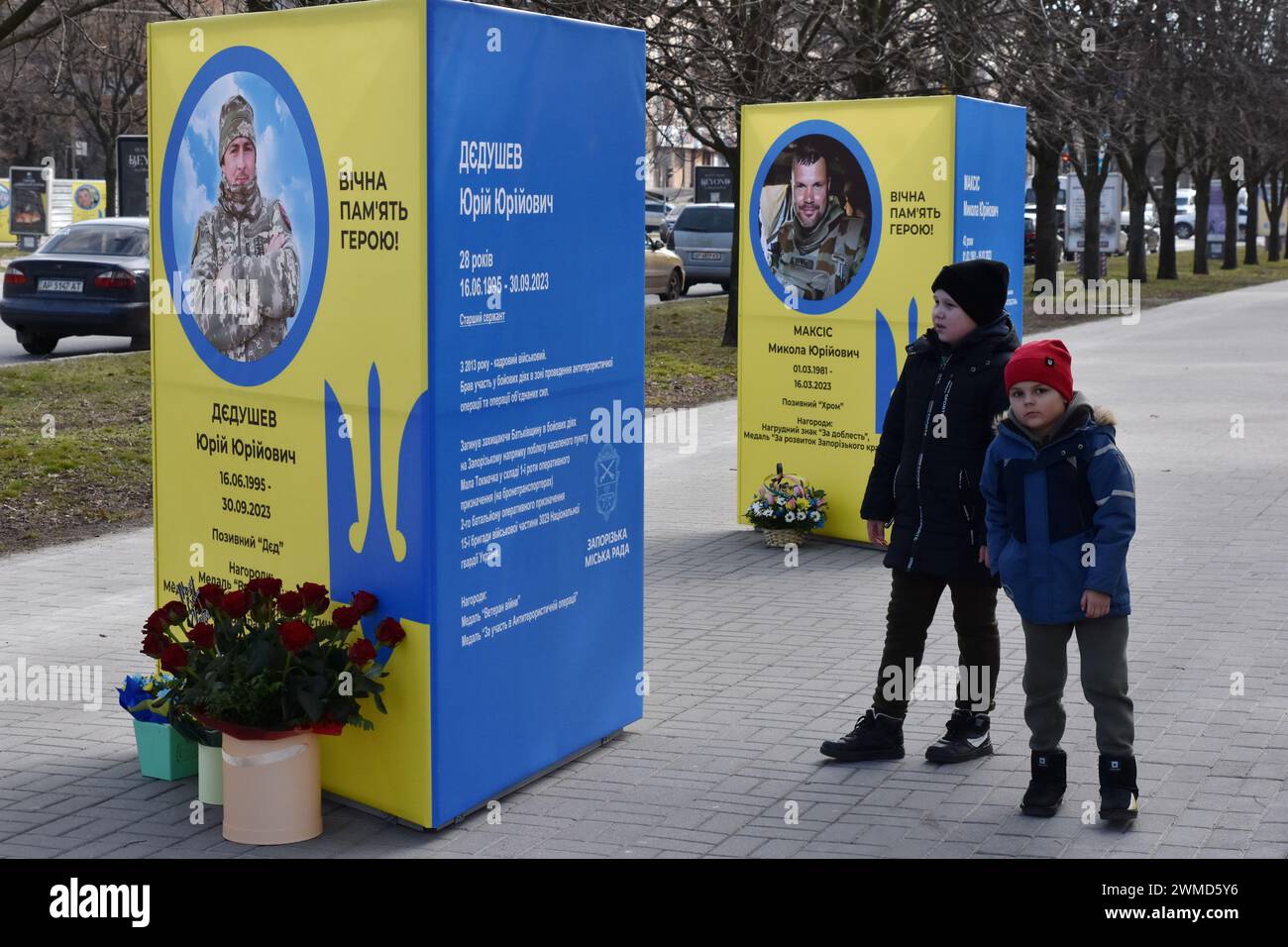 De jeunes garçons sont vus à côté des stands de portraits de soldats ukrainiens tombés au combat dans le centre de Zaporizhzhia. Le président ukrainien Volodymyr Zelensky a marqué le deuxième anniversaire de l'invasion russe en déclarant que son pays "réussira". L'Ukraine a défié les prédictions de combattre la Russie, mais il y a des signes que la guerre tourne en faveur de la Russie. Le gouvernement ukrainien a cessé de partager le nombre de ses militaires morts il y a longtemps. Mais le nombre toujours croissant de tombes fraîches, des deux côtés des lignes de front, dresse un tableau douloureusement clair. Banque D'Images