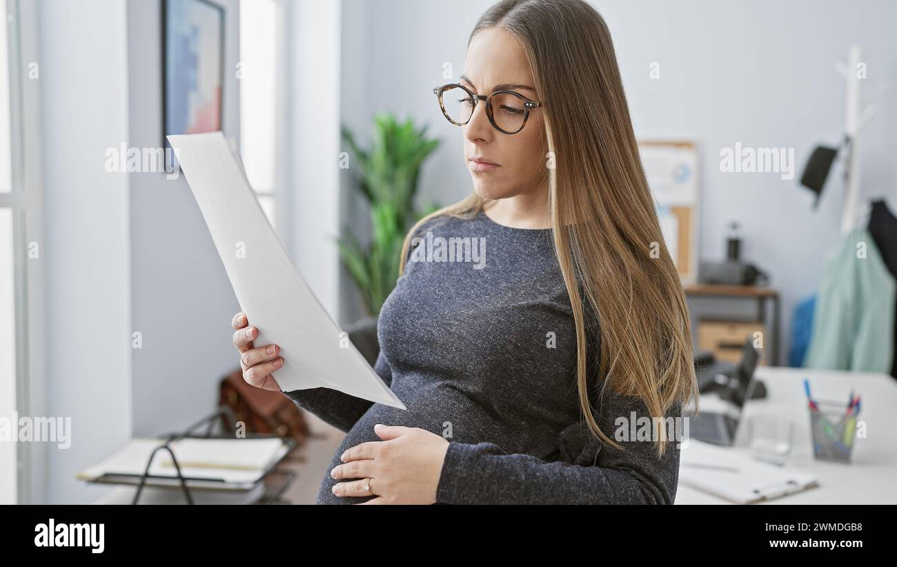 Une femme hispanique enceinte lit un document dans un bureau, décrivant la maternité sur le lieu de travail. Banque D'Images