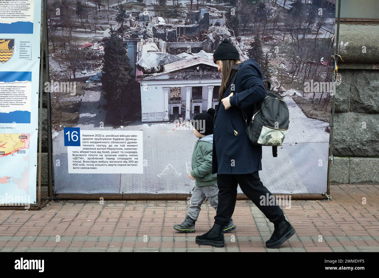 Kiev, Ukraine. 24 février 2024. Une femme et un enfant marchent près d'une photo du théâtre dramatique Marioupol détruit par une frappe aérienne russe, dans le centre de Kiev. Alors que l'Ukraine marque deux ans depuis l'invasion russe à grande échelle, une humeur sombre plane sur le pays. Sur le champ de bataille, les troupes ukrainiennes manquent de munitions alors qu’elles cherchent une aide occidentale supplémentaire. Crédit : SOPA images Limited/Alamy Live News Banque D'Images