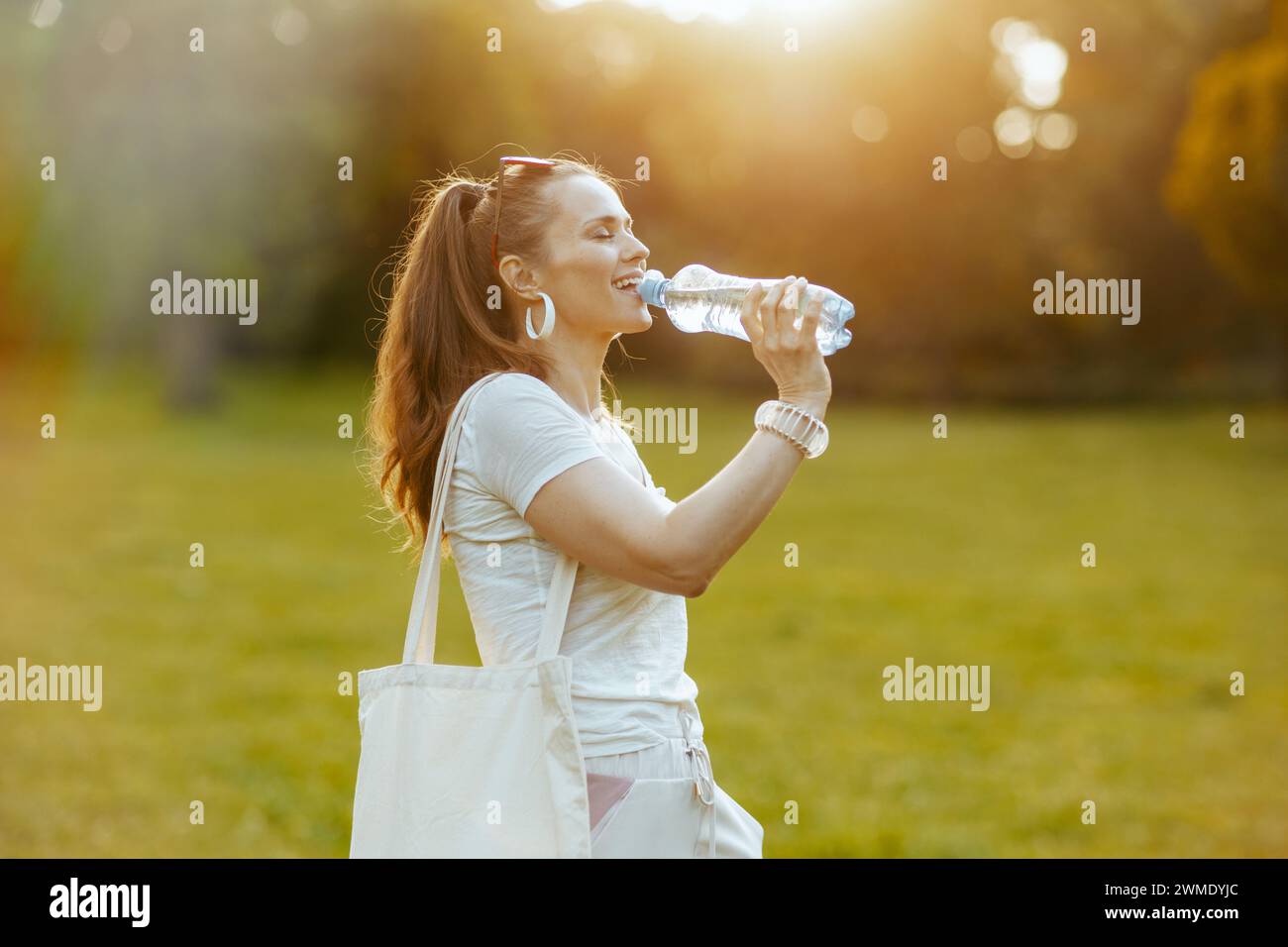 Heure d'été. décontracté tendance femme de 40 ans en chemise blanche avec sac fourre-tout et bouteille d'eau dans la prairie à l'extérieur. Banque D'Images