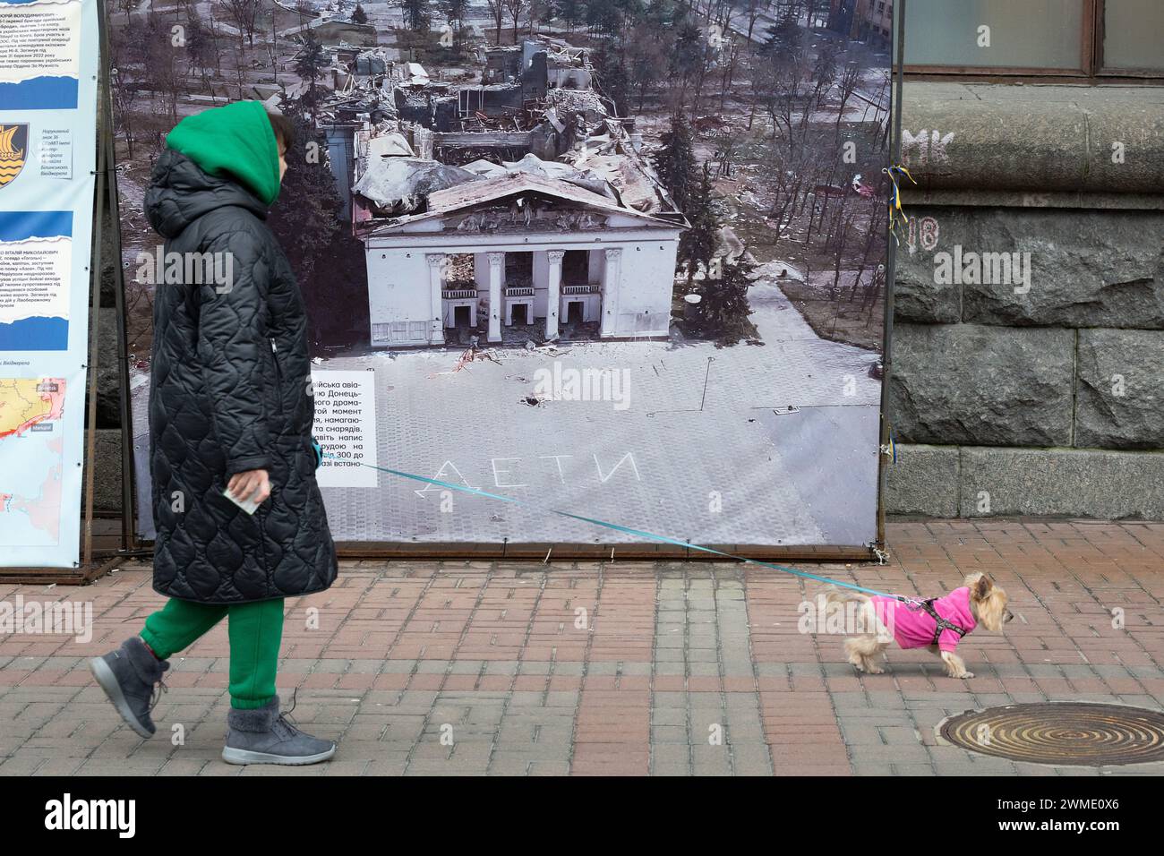 Kiev, Ukraine. 24 février 2024. Une femme marche avec son chien près d'une photo du théâtre dramatique Marioupol détruit par une frappe aérienne russe, dans le centre de Kiev. Alors que l'Ukraine marque deux ans depuis l'invasion russe à grande échelle, une humeur sombre plane sur le pays. Sur le champ de bataille, les troupes ukrainiennes manquent de munitions alors qu’elles cherchent une aide occidentale supplémentaire. (Photo de Oleksii Chumachenko/SOPA images/SIPA USA) crédit : SIPA USA/Alamy Live News Banque D'Images