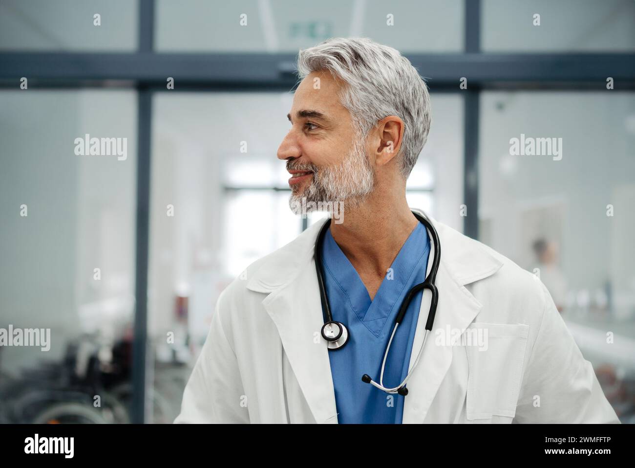 Portrait d'un médecin adulte confiant debout dans le couloir de l'hôpital. Beau médecin avec les cheveux gris portant un manteau blanc, gommages, stéthoscope autour du cou Banque D'Images