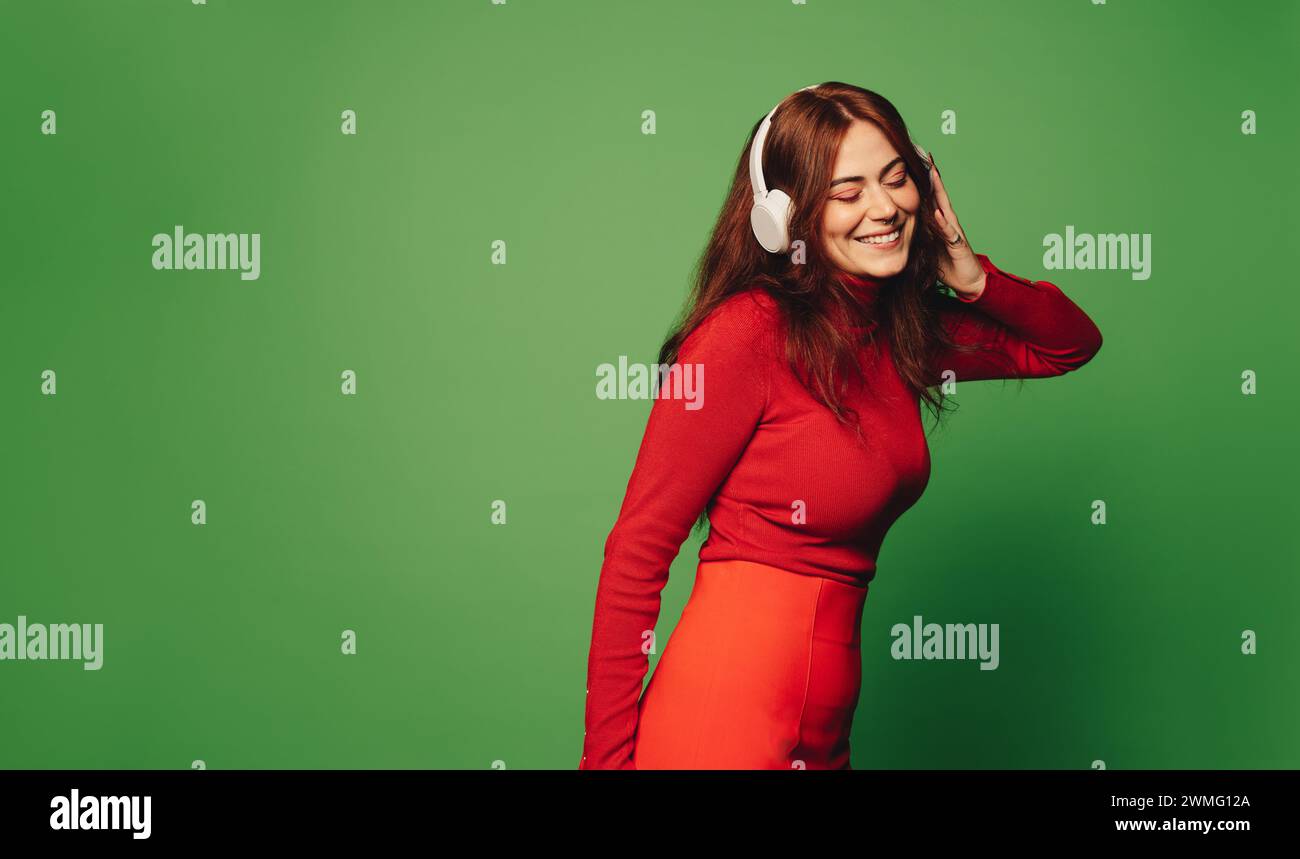 Femme joyeuse dans des vêtements décontractés à la mode est debout dans un studio vert vibrant, écoutant de la musique sur des écouteurs sans fil et souriant. Animateurs Banque D'Images