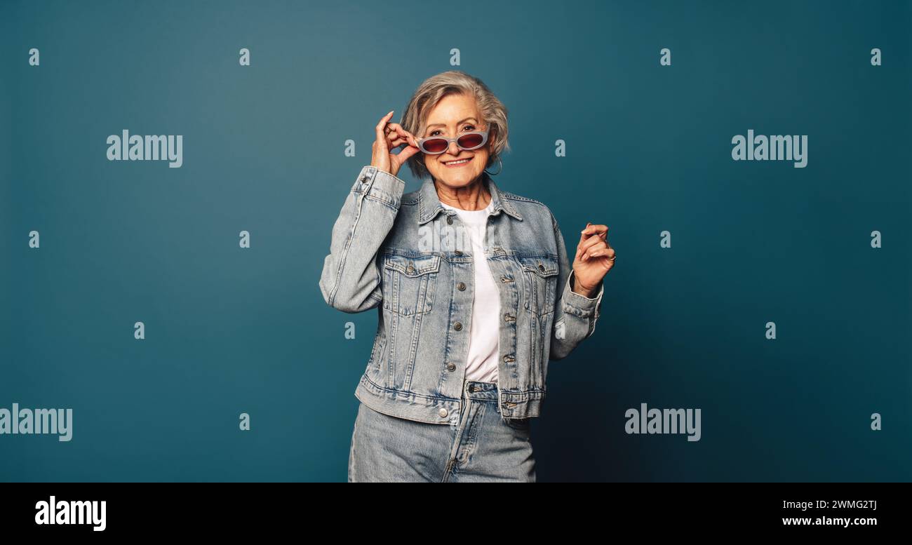 Femme âgée joyeuse dans une tenue décontractée en denim et des lunettes élégantes se tient ludique dans un studio. Sa personnalité insouciante et amusante brille à travers un Banque D'Images