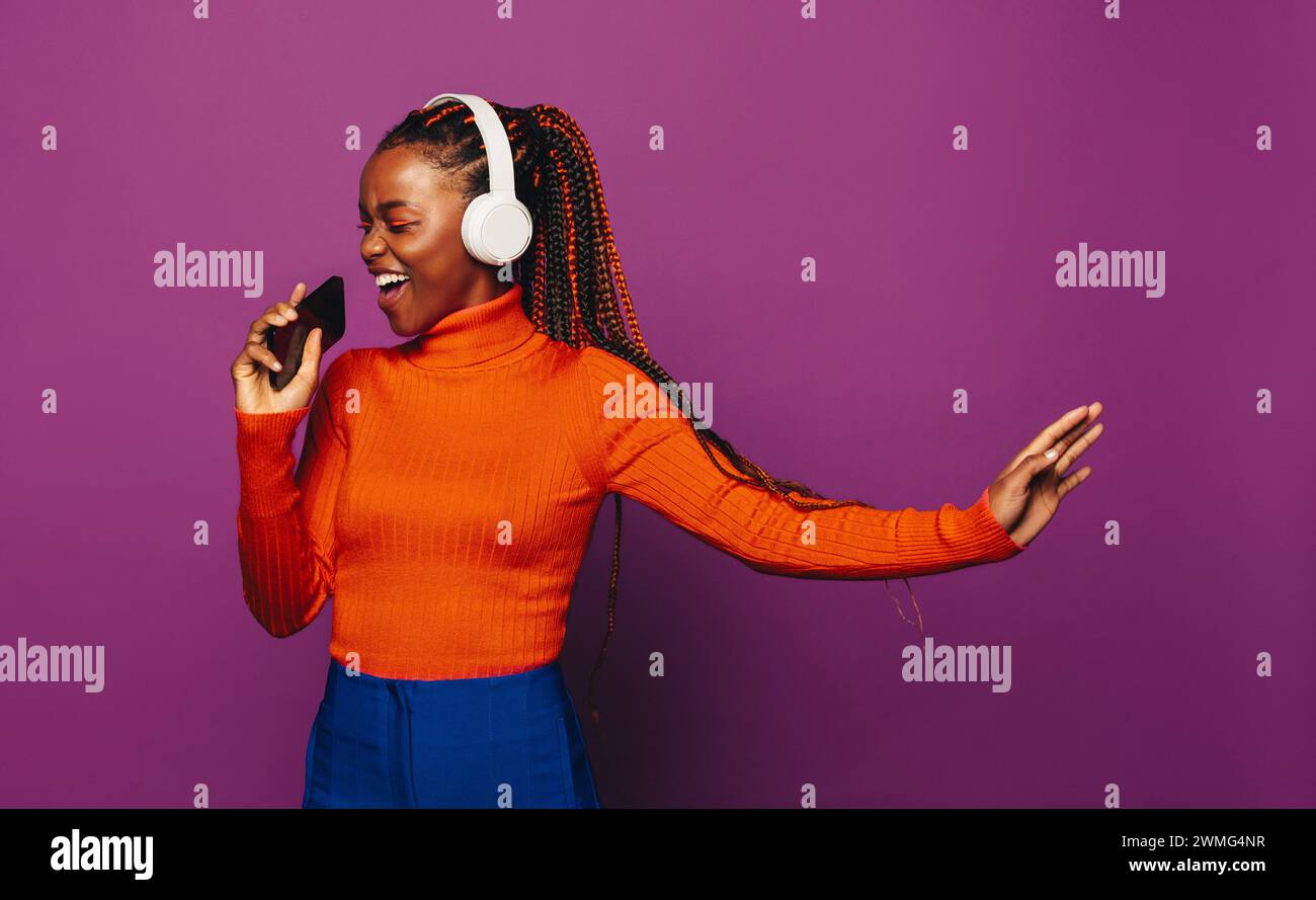 Femme africaine joyeuse avec des tresses bicolores se tient dans un studio vibrant, tenant un smartphone sur un fond violet. Elle écoute de la musique avec lui Banque D'Images