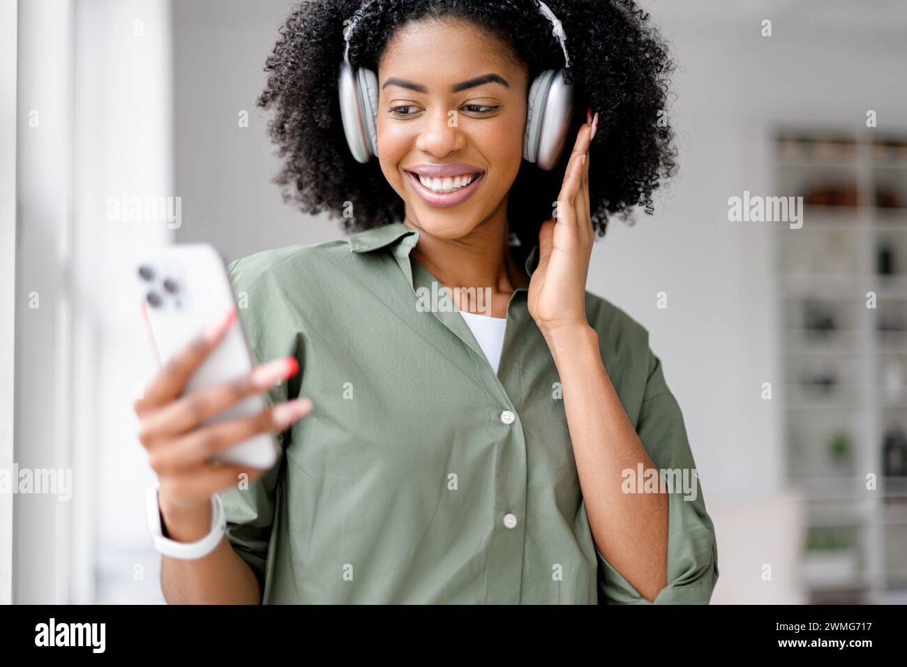Une femme afro-américaine vivante portant des écouteurs profite d'un appel vidéo animé, son sourire illuminé par la lumière du jour qui traverse une fenêtre dans un cadre de bureau chic et minimaliste. Banque D'Images