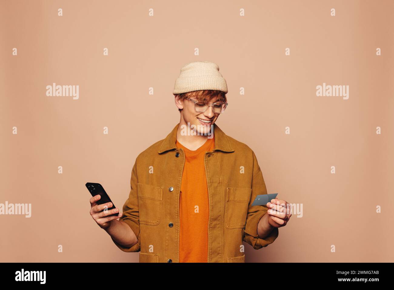 Heureux jeune homme en veste denim décontractée tient une carte de crédit et un téléphone portable sur un fond de pêche vibrant. Représentant les services bancaires mobiles, les achats en ligne Banque D'Images