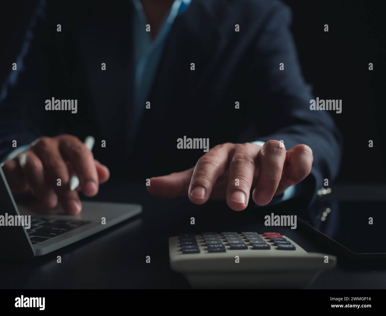 Homme d'affaires en costume noir travaillant sur ordinateur portable et tablette et calculatrice, mains toucher sur tablette au bureau avec fond sombre, travail en ligne Banque D'Images