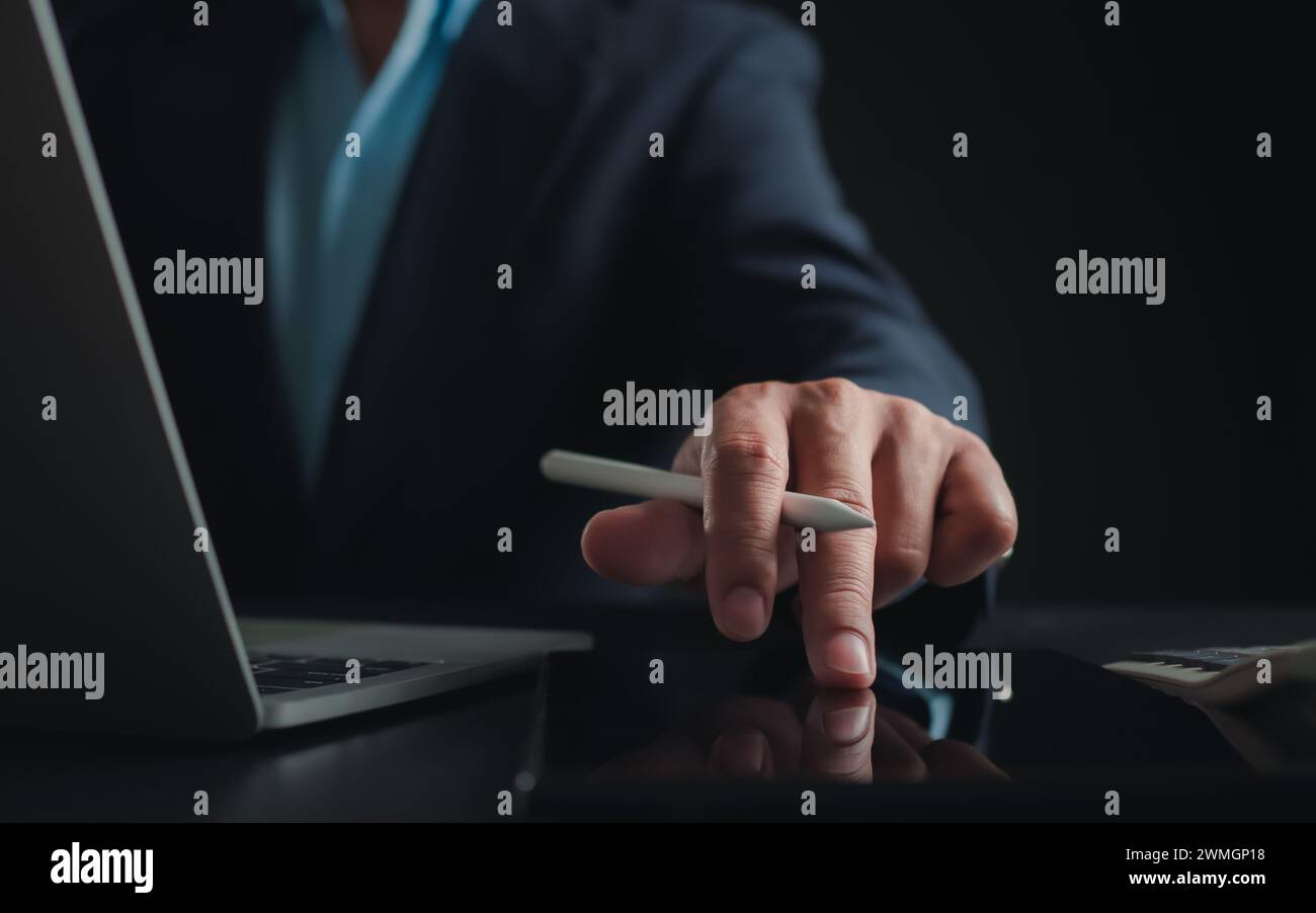 Homme d'affaires en costume noir travaillant sur ordinateur portable et tablette, mains toucher sur tablette au bureau avec fond sombre, travail en ligne, gros plan, Banque D'Images
