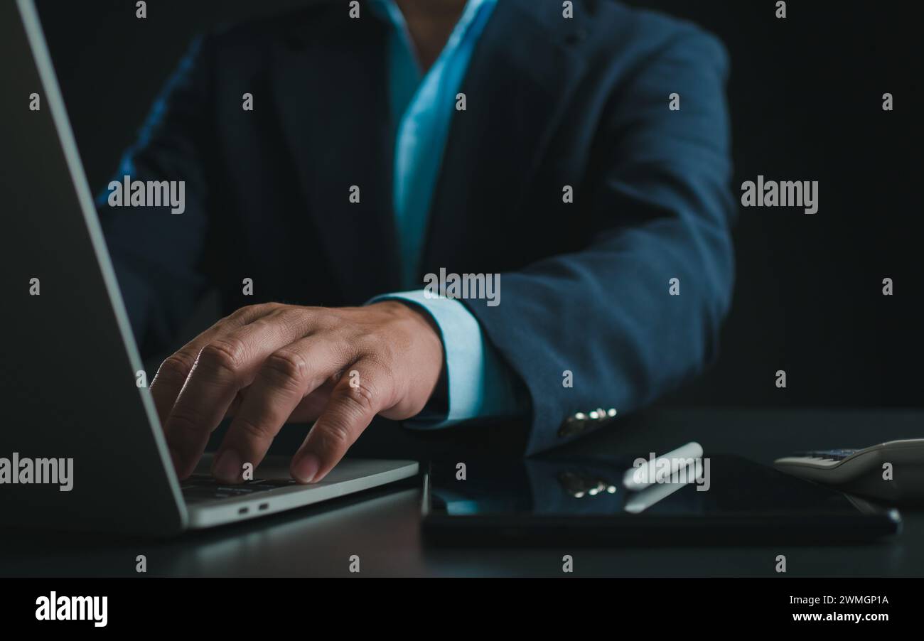Homme d'affaires en costume noir travaillant sur ordinateur portable et tablette, tapant à la main sur le clavier sur la table au bureau avec fond sombre, travail en ligne, Clos Banque D'Images