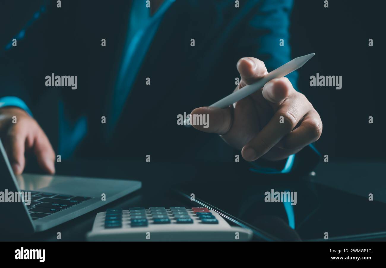 Homme d'affaires en costume noir travaillant sur ordinateur portable et tablette et calculatrice, mains toucher sur tablette au bureau avec fond sombre, travail en ligne Banque D'Images