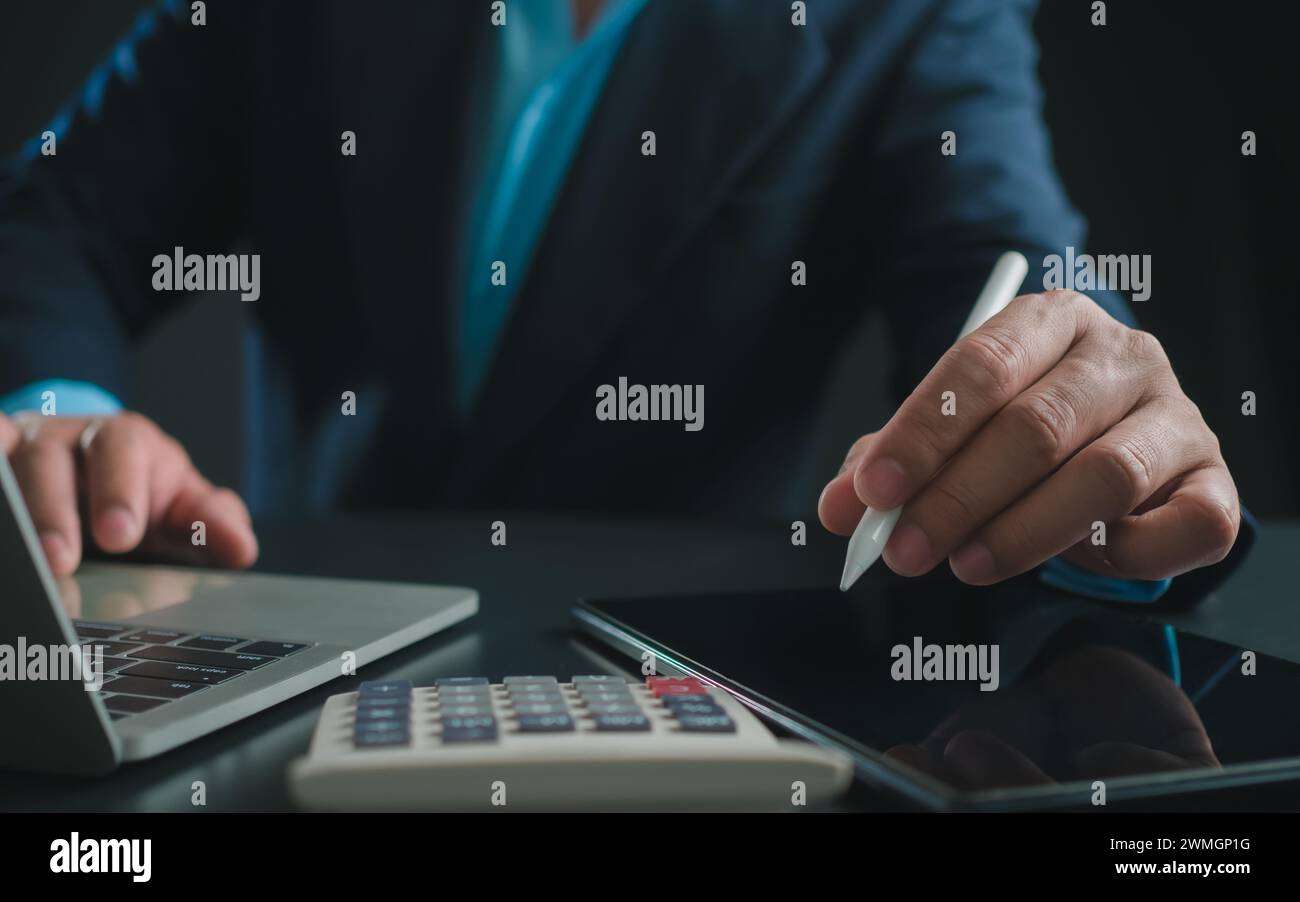 Homme d'affaires en costume noir travaillant sur ordinateur portable et tablette et calculatrice, mains toucher sur tablette au bureau avec fond sombre, travail en ligne Banque D'Images