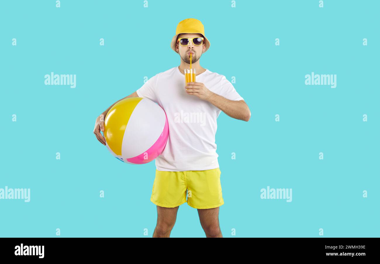 Vacancier masculin drôle avec boule de plage gonflable buvant cocktail orange sur fond bleu clair. Banque D'Images