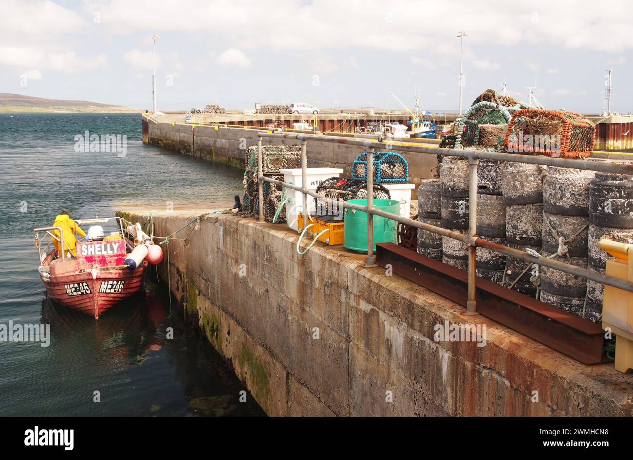 Un petit bateau de pêche avec un homme à côté de la jetée de Tingwall, avec des piles de nasses de pêche et des boîtes sur la jetée et d'autres bateaux amarrés, Orcades, Écosse, Royaume-Uni Banque D'Images