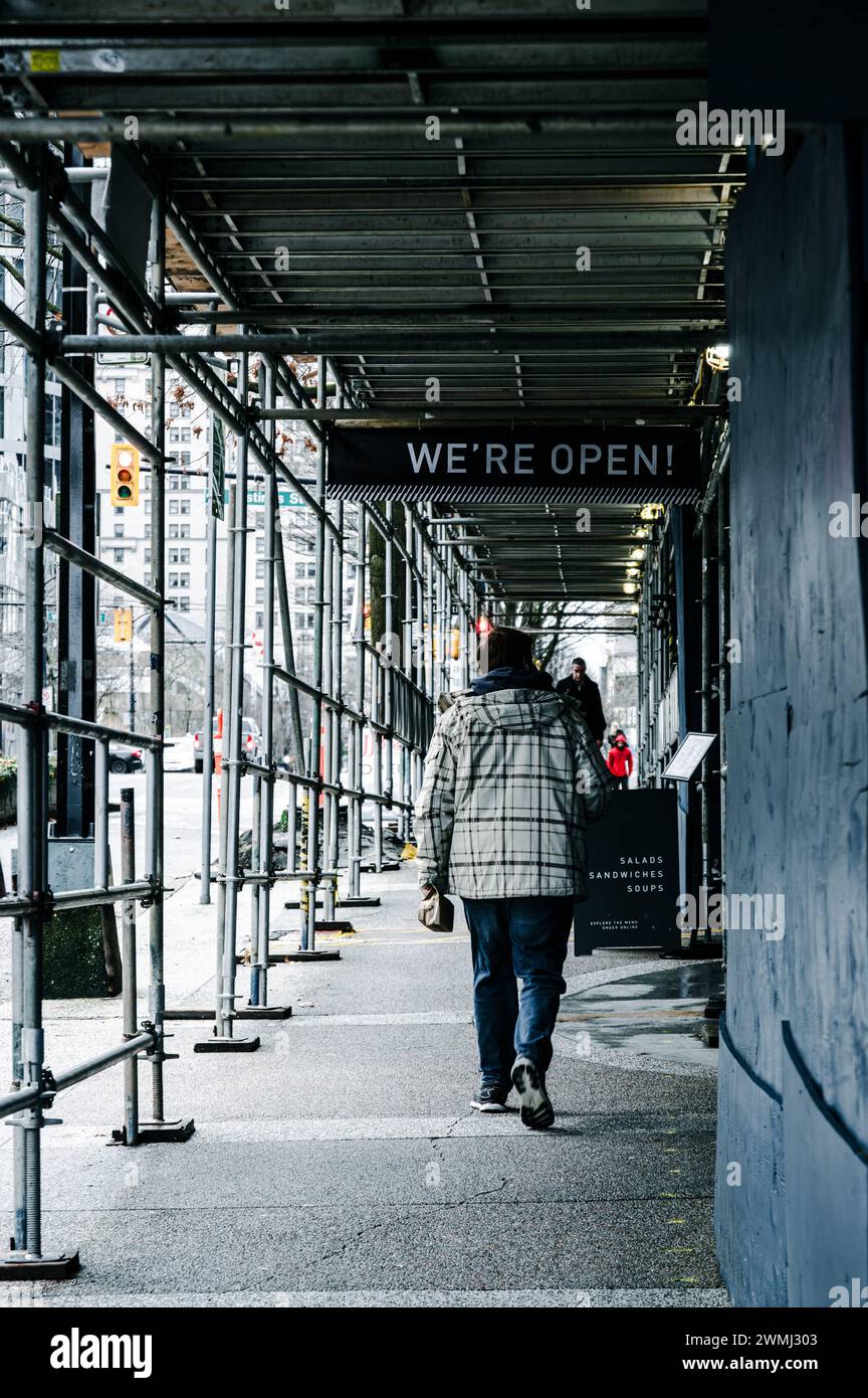 Un homme marchant le long du trottoir de Burrard Street, sous un échafaudage élevé pour des travaux de construction. Un panneau indique qu'un café est encore ouvert. Banque D'Images