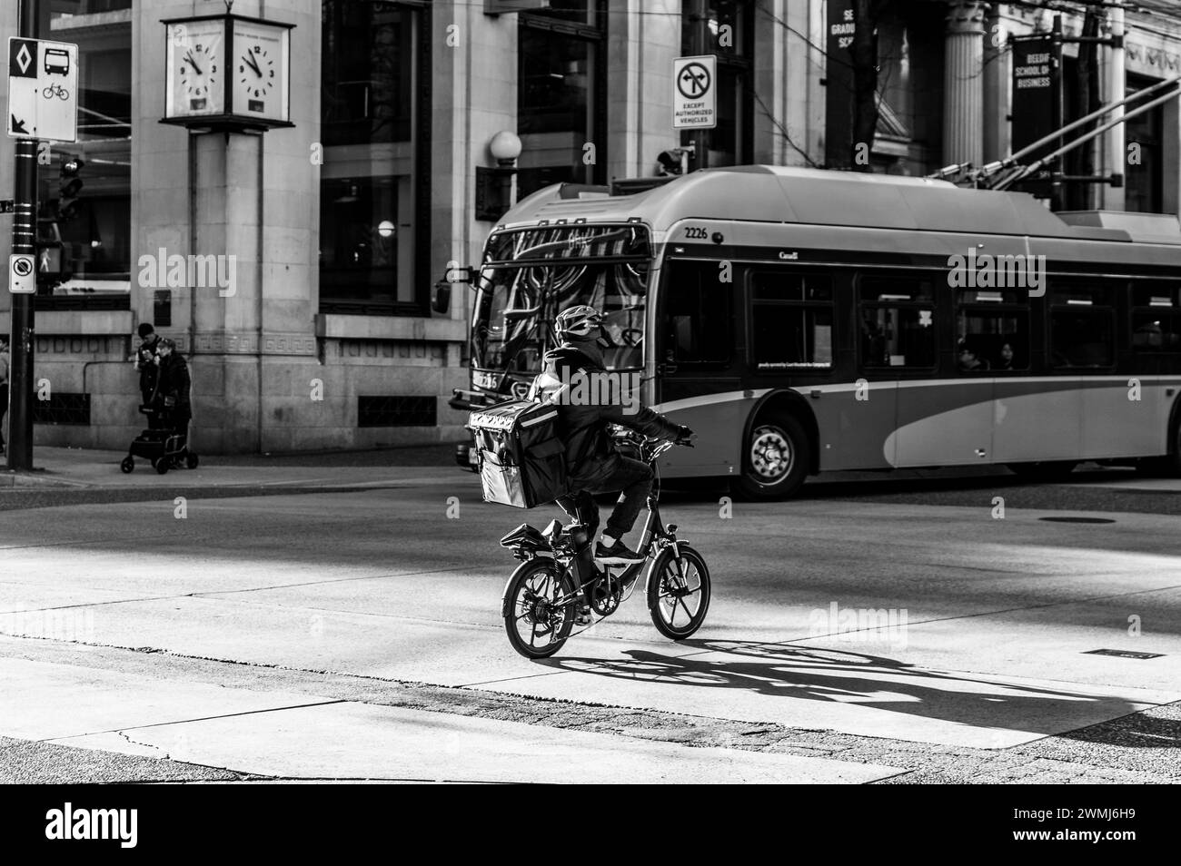 Vancouver, Canada - 16 févr. 2024 : photo en noir et blanc d'un homme chevauchant son vélo électrique et traversant la rue Pender, avec un trolleybus tournant. Banque D'Images