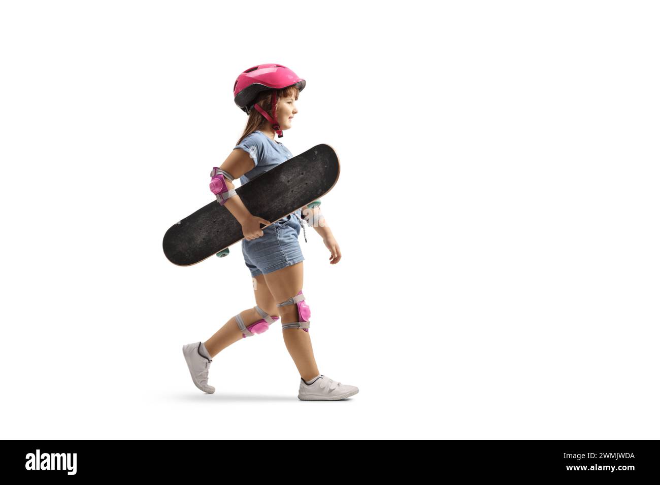 Plan de profil pleine longueur d'une petite fille marchant et portant une planche à roulettes, portant un coude, des genouillères et un casque isolé sur fond blanc Banque D'Images