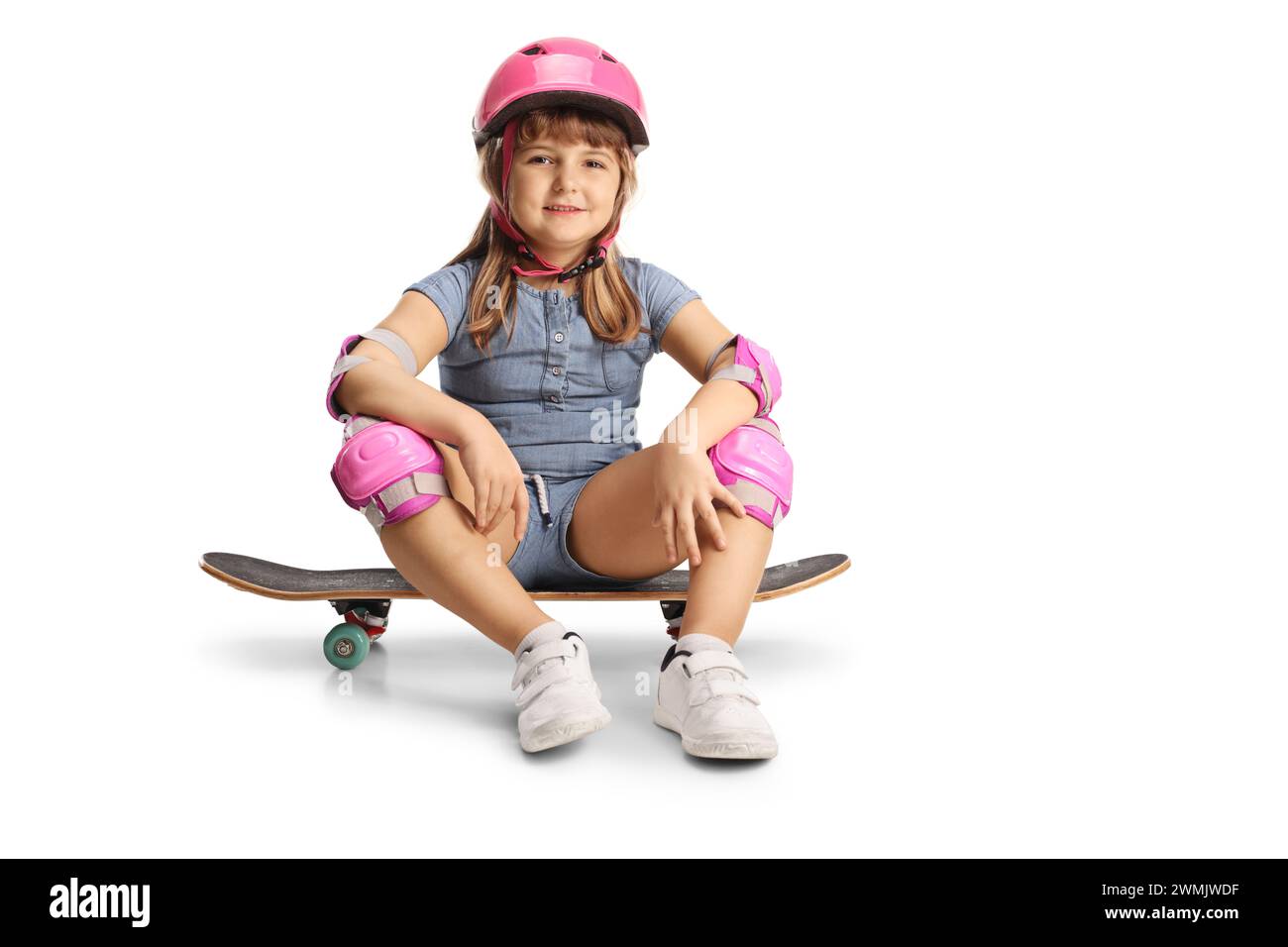 Plan de profil pleine longueur d'une petite fille assise sur une planche à roulettes, portant un coude, des genouillères et un casque isolé sur fond blanc Banque D'Images