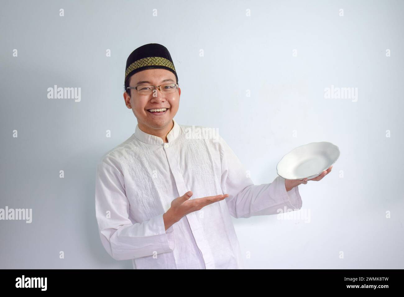 Homme musulman asiatique portant des lunettes et un tissu blanc pointant la plaque vide pour jeûner ramadhan. Fond blanc isolé Banque D'Images