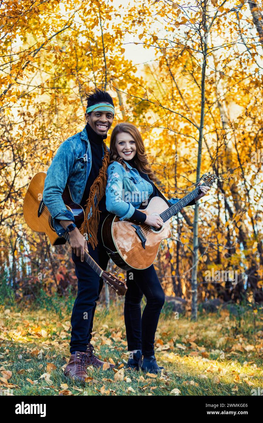 Couple marié de race mixte, souriant et posant pour la caméra tenant des guitares lors d'une sortie en famille d'automne dans un parc de la ville, passer du temps de qualité toge... Banque D'Images