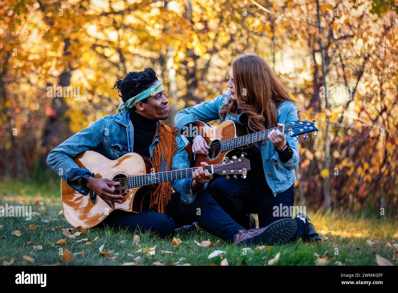 Couple marié de race mixte assis sur l'herbe se regardant, passant du temps de qualité ensemble à jouer des guitares lors d'une sortie en famille d'automne dans un c... Banque D'Images