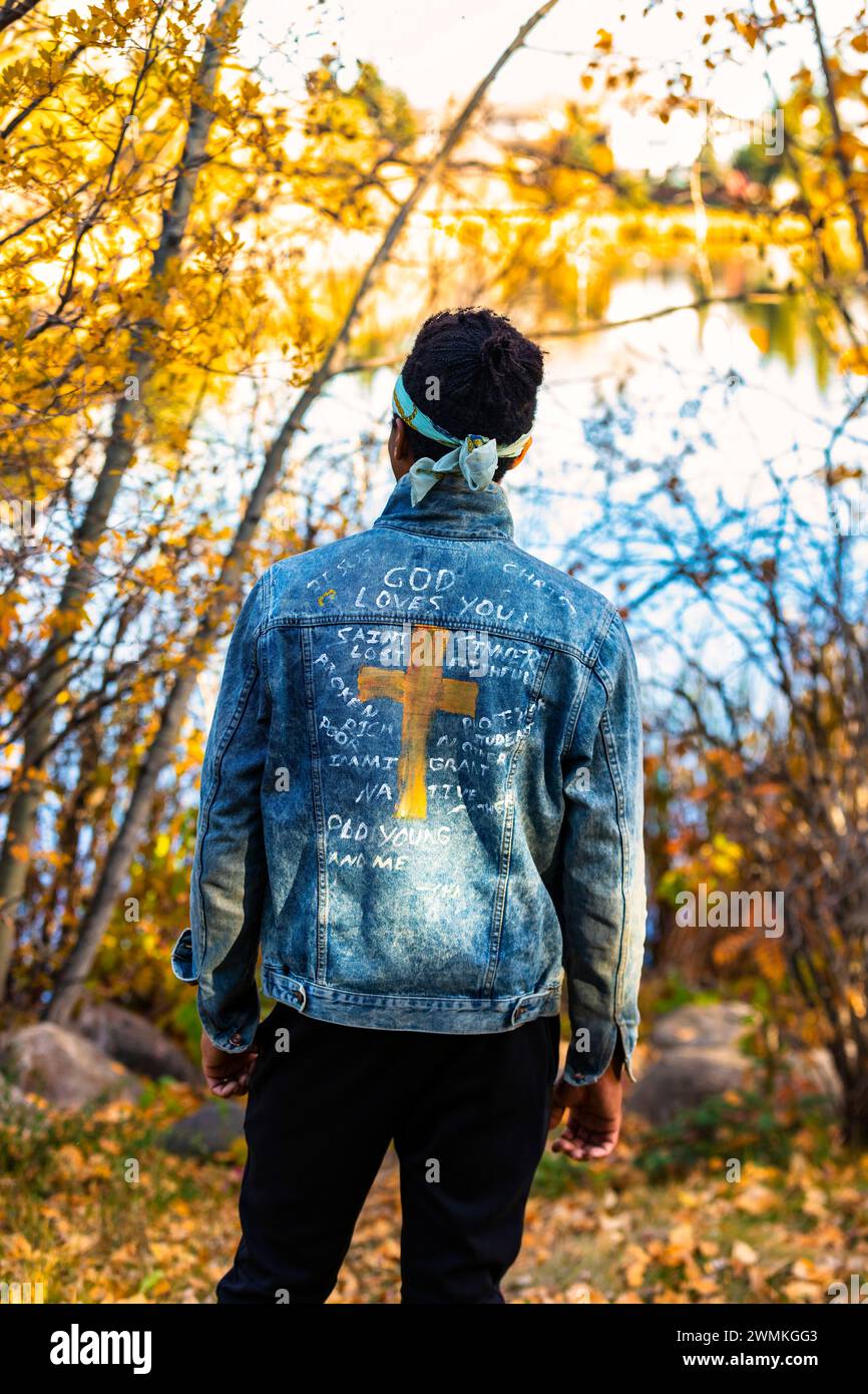 Vue rapprochée de derrière un homme portant une veste en jean avec des références chrétiennes manuscrites lors d'une sortie d'automne dans un parc de la ville, debout près d'un ... Banque D'Images